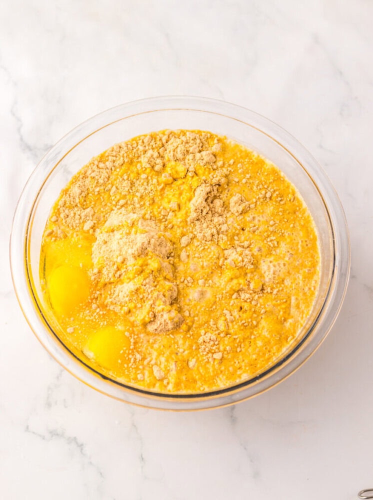 A glass mixing bowl containing raw eggs, dry cake mix, and liquid ingredients on a white marble surface.