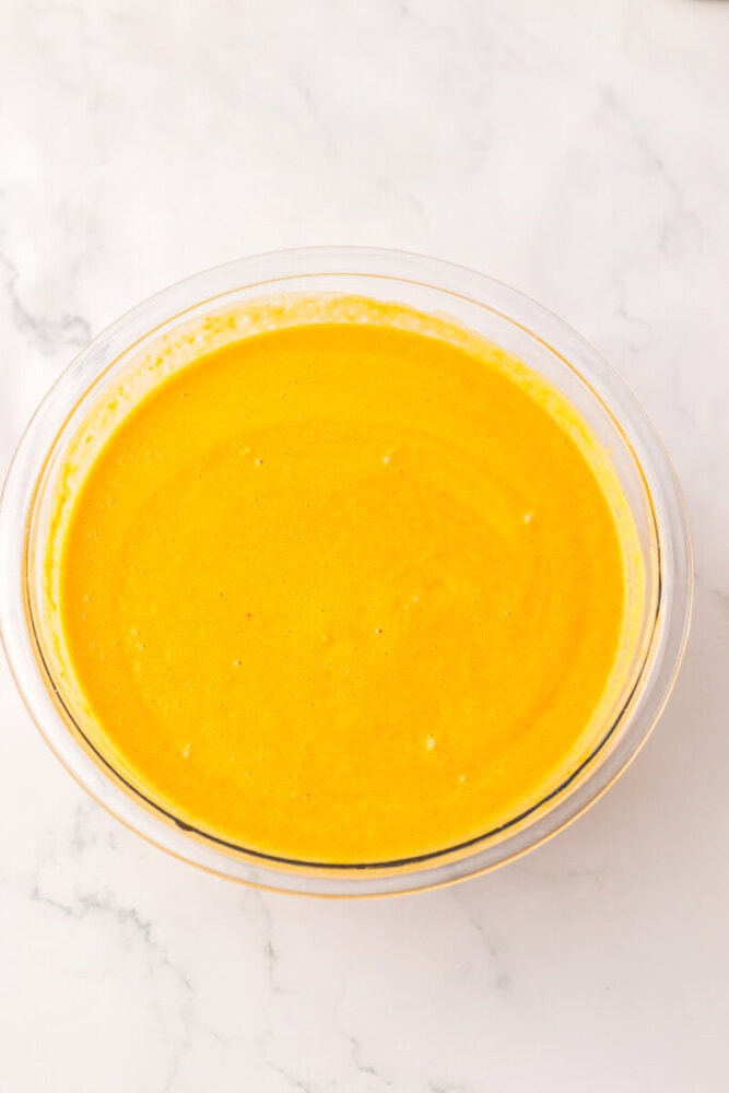 A clear glass bowl filled with a smooth, bright yellow liquid mixture sits on a white marble surface.