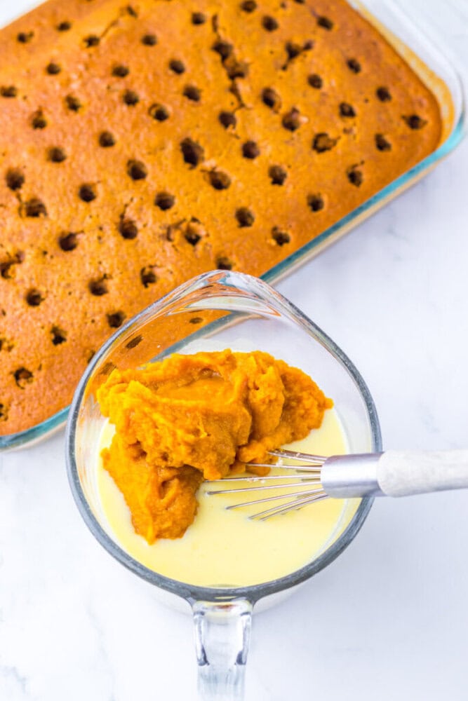 A glass dish of poke cake with holes on top is next to a mixing bowl containing pumpkin puree and a whisk in a creamy mixture.