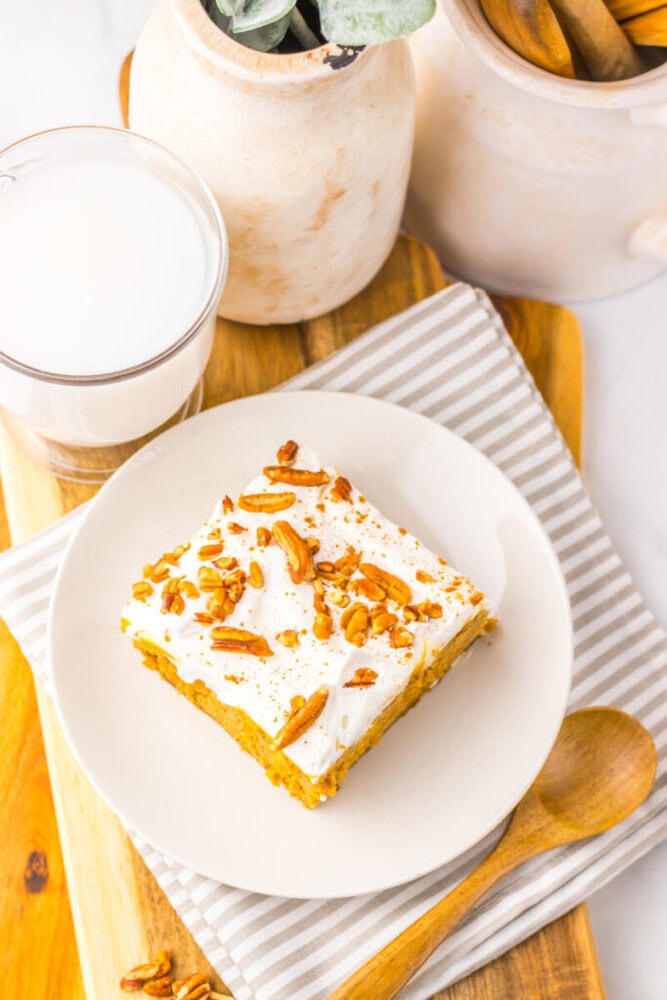 A slice of frosted cake topped with chopped nuts sits on a white plate next to a glass of milk and a wooden spoon on a striped napkin.