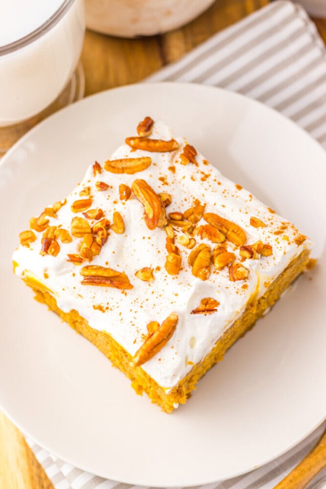 A square slice of cake with white frosting and chopped pecans on top, served on a white plate.