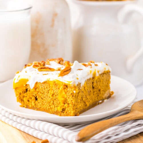 A slice of pumpkin cake with white frosting and pecans on a white plate, next to a glass of milk and a wooden spoon.