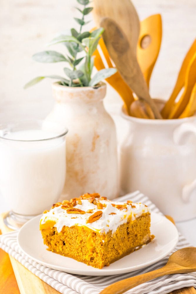 A slice of frosted pumpkin cake with chopped pecans on a white plate, next to a glass of milk; wooden utensils and a plant in the background.