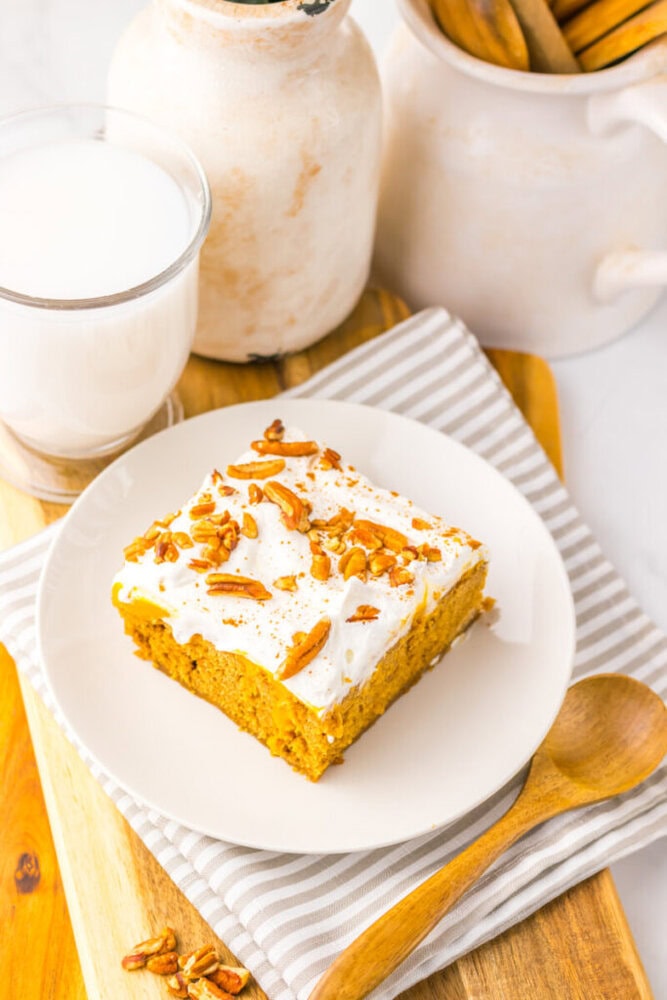A slice of pumpkin cake with white frosting and chopped pecans on a white plate, next to a glass of milk and a wooden spoon on a striped napkin.