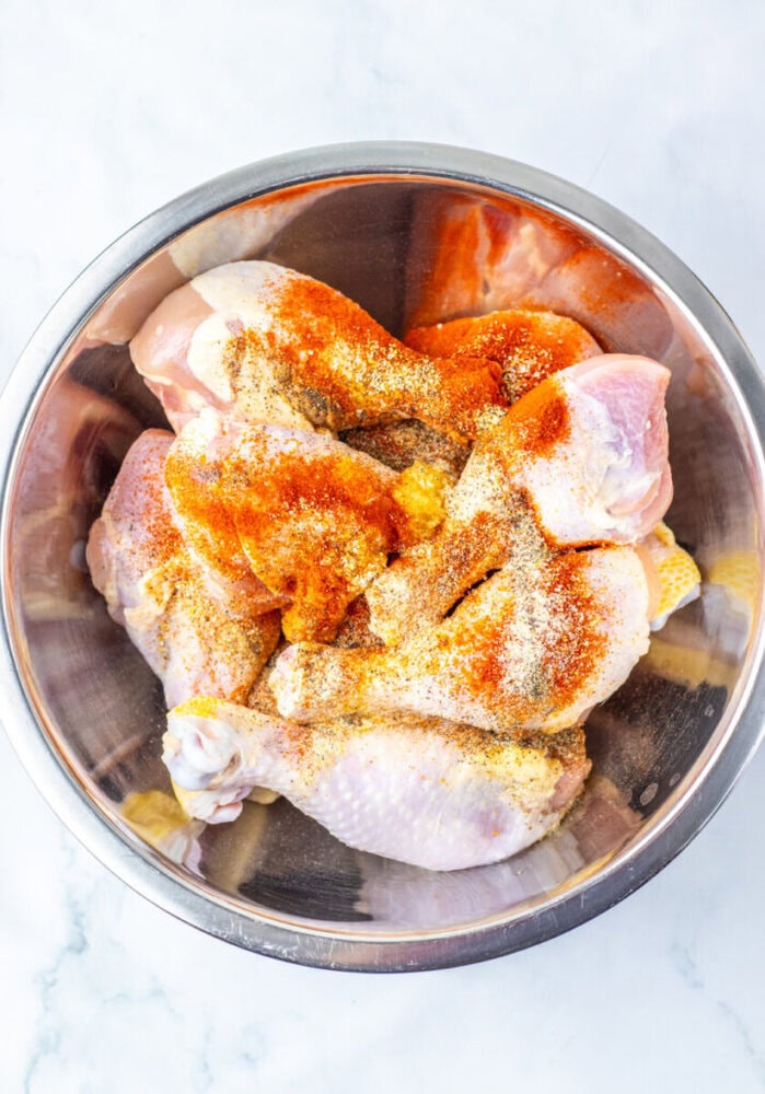 Raw chicken drumsticks in a metal bowl, topped with various spices including paprika and garlic powder, on a light marble surface.