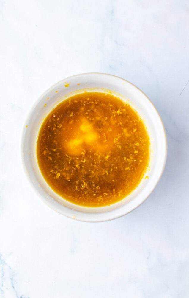 A white bowl filled with a yellow-orange liquid mixture, likely a sauce or dressing, on a white marble surface.