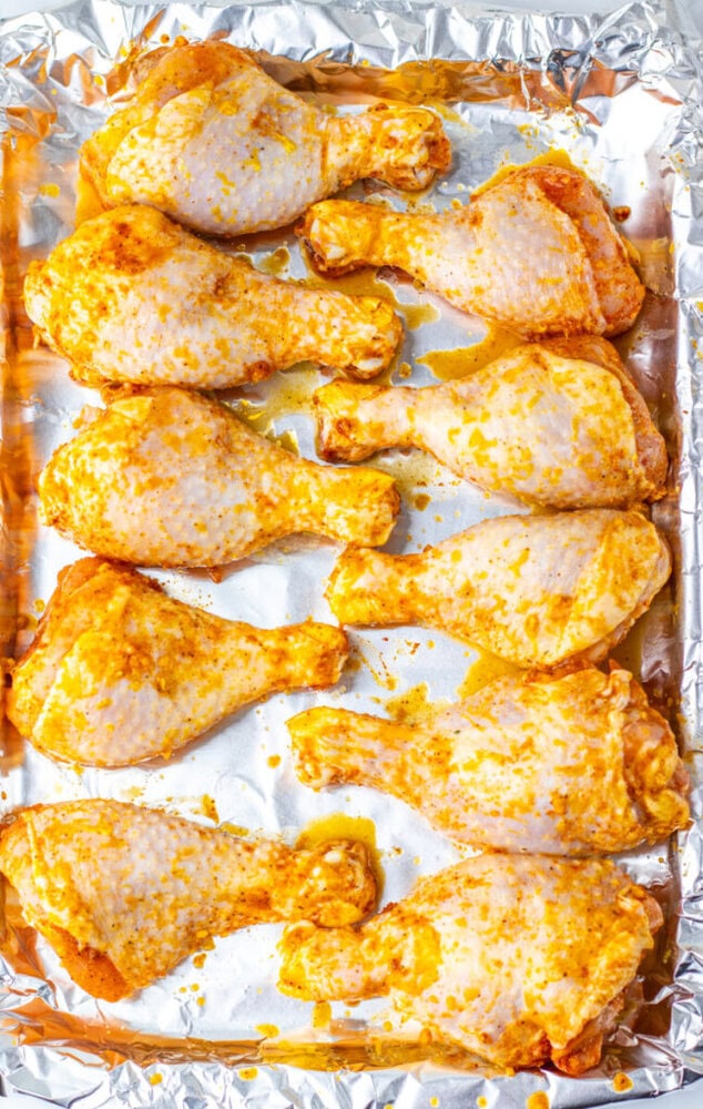 Ten raw, seasoned chicken drumsticks arranged on a foil-lined baking sheet, ready to be cooked.