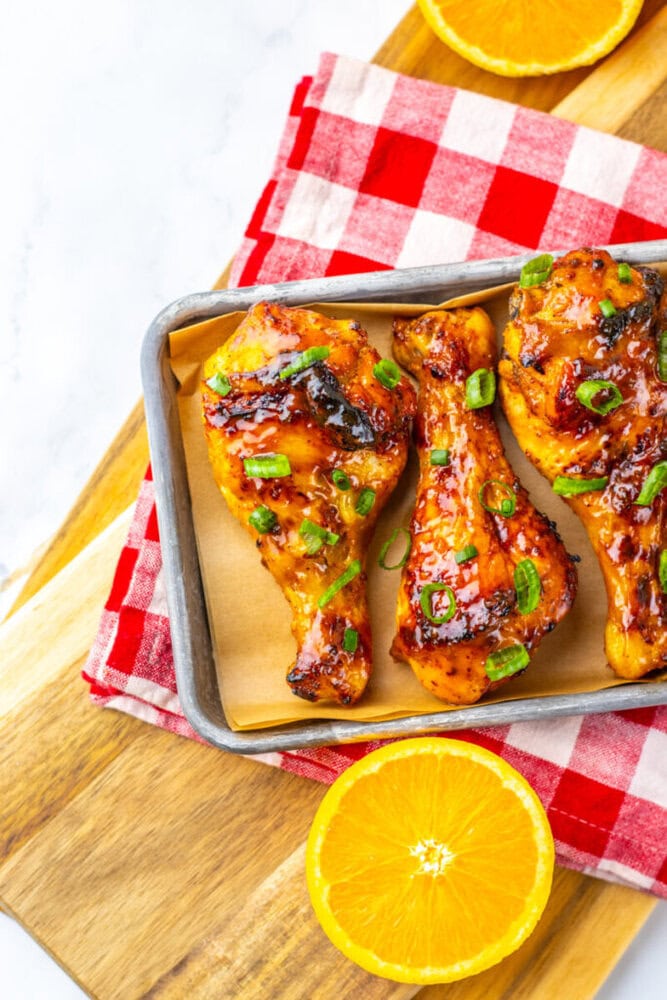 Baked chicken drumsticks garnished with green onions on parchment in a tray, set on a red checkered napkin, with orange slices on a wooden board.