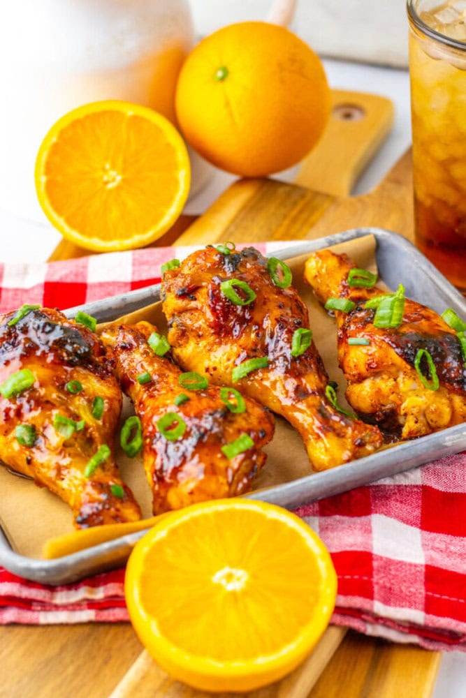 Three pieces of glazed chicken garnished with green onions on a tray, with halved oranges and a glass of iced tea in the background.