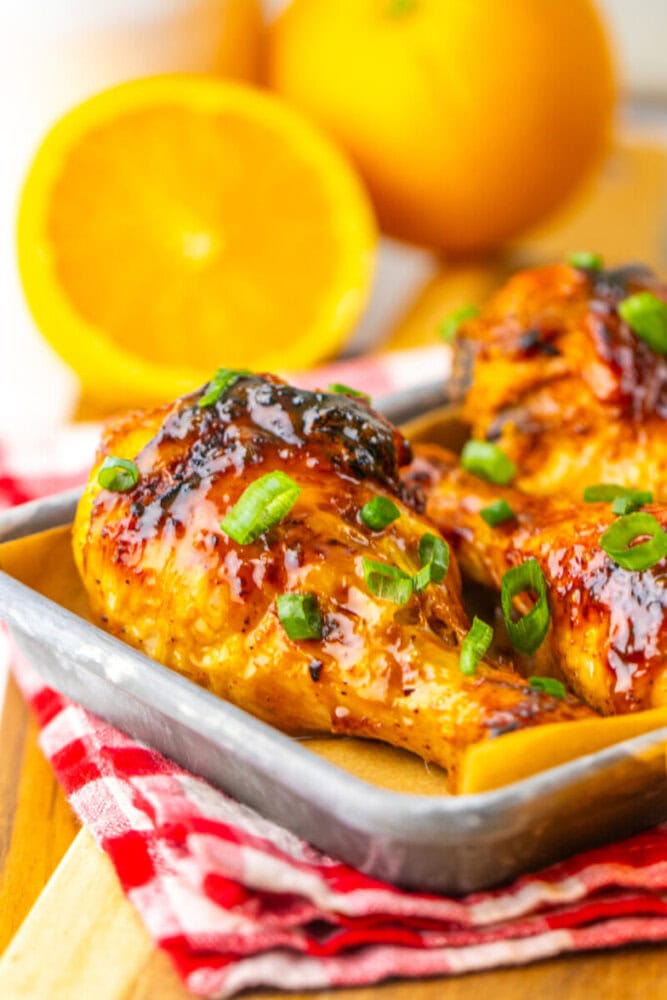 Baked chicken legs glazed with sauce and garnished with sliced green onions, served on a metal tray with oranges in the background.