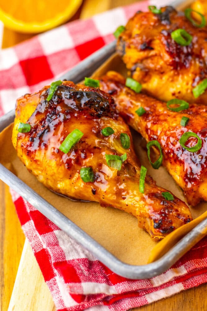 Baked chicken drumsticks glazed with sauce and garnished with chopped green onions on a tray, set on a red and white checkered cloth.