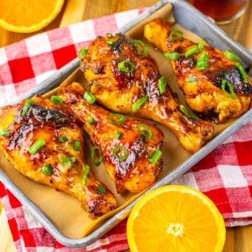 Four glazed chicken drumsticks garnished with chopped green onions on parchment paper in a tray, with orange slices and a red checkered cloth underneath.