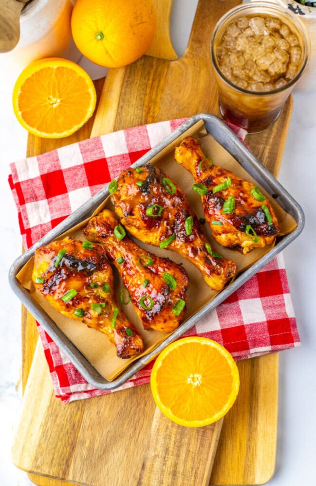 Three glazed chicken drumsticks garnished with green onions on a tray, with orange slices, a drink with ice, and a red gingham napkin on a wooden board.