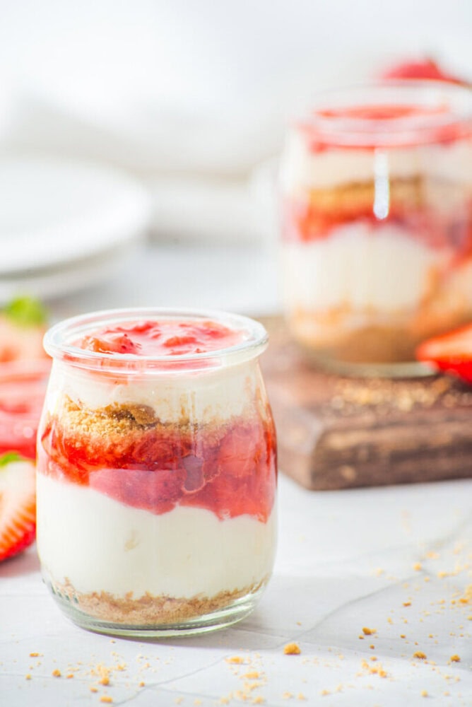 A glass jar filled with layered cheesecake, strawberry sauce, and graham cracker crumbs sits on a white surface; another jar is in the background.