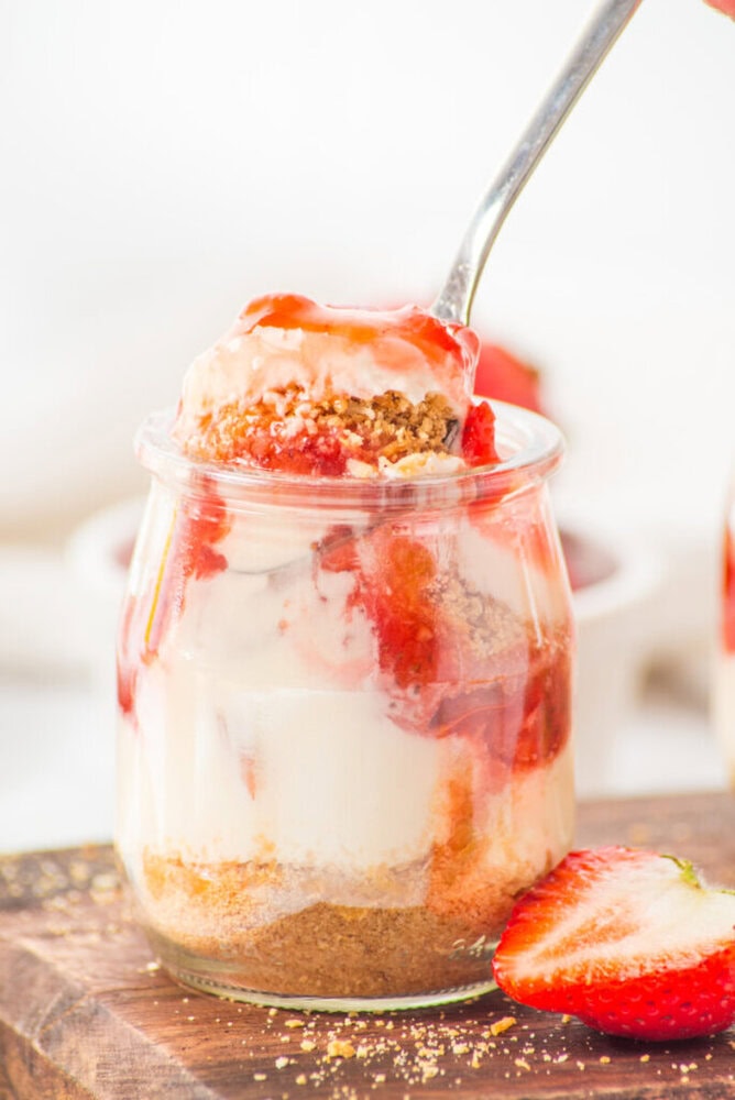 A glass jar filled with layered cheesecake, strawberry sauce, and graham cracker crumbs, with a spoon scooping out a portion. A sliced strawberry lies beside the jar.