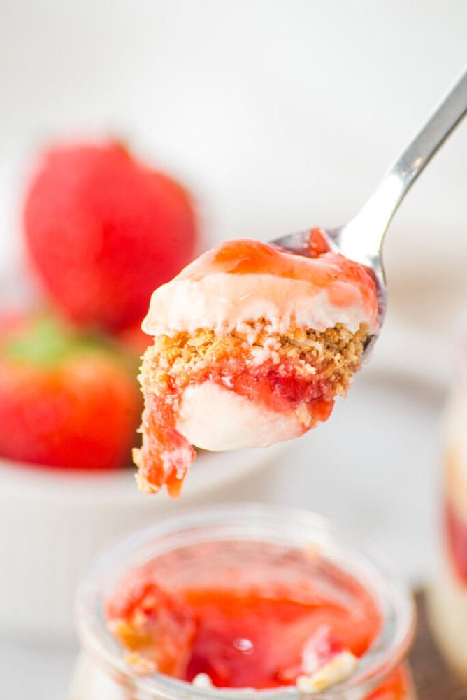A spoon holds a layered dessert with whipped cream, strawberry topping, and a crumbly crust, with fresh strawberries blurred in the background.