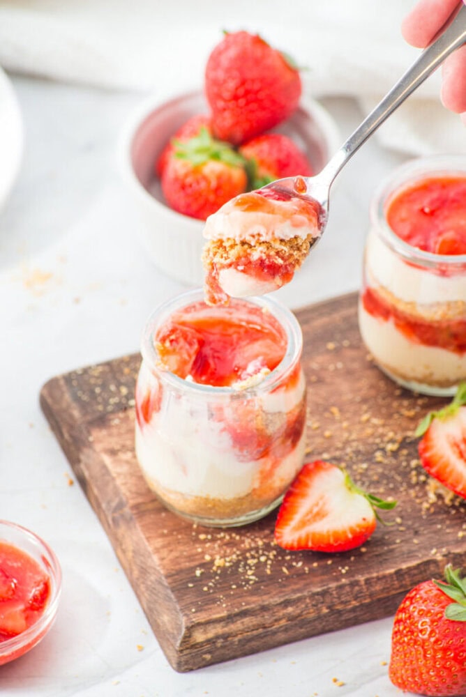 A spoon holds a portion of layered strawberry cheesecake in a jar, with fresh strawberries and more cheesecake jars on a wooden board.
