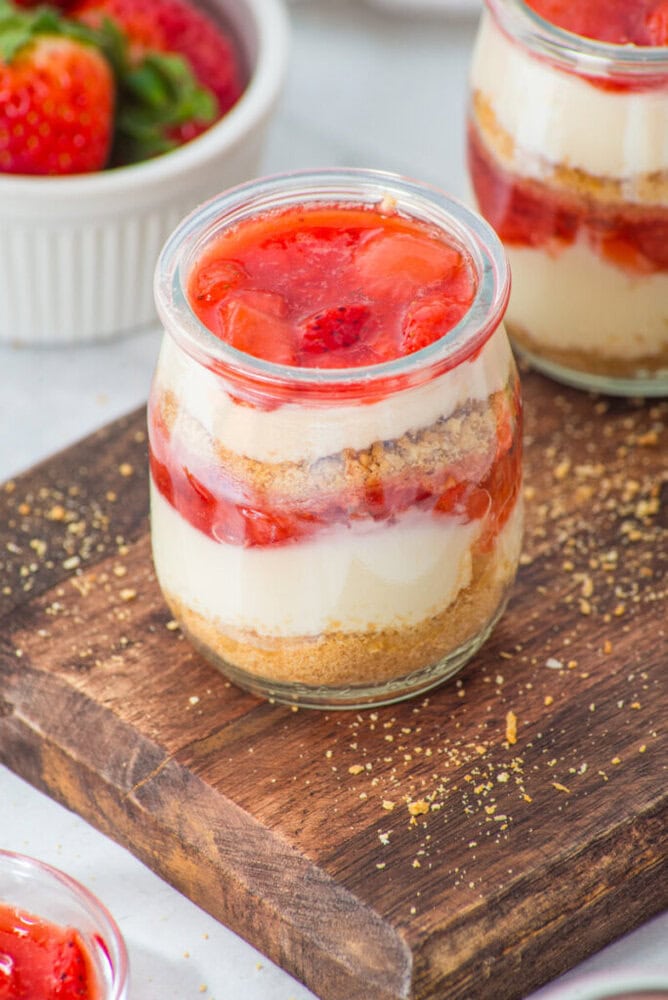 A glass jar filled with layers of cheesecake, graham cracker crumbs, and strawberry topping sits on a wooden board.