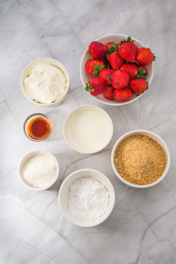 Bowls containing strawberries, cream cheese, heavy cream, graham cracker crumbs, powdered sugar, shredded coconut, and vanilla extract on a marble surface.