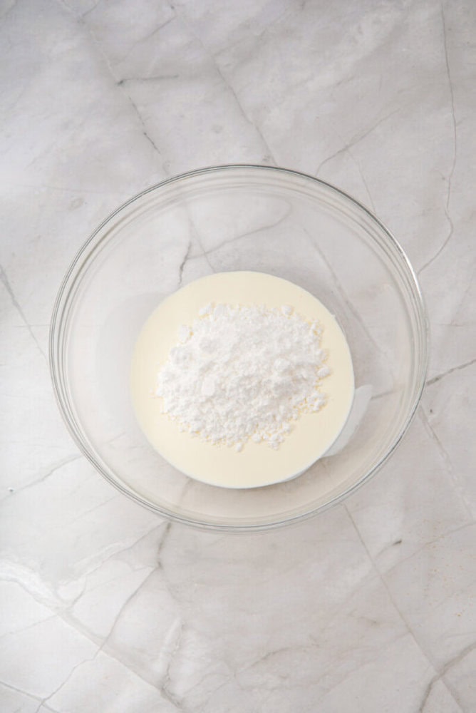 A clear glass bowl containing heavy cream and powdered sugar sits on a white marble surface.