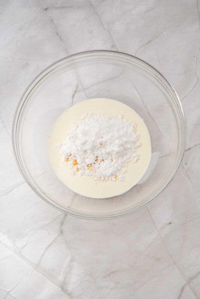 A glass bowl containing heavy cream and powdered sugar sits on a marble surface.