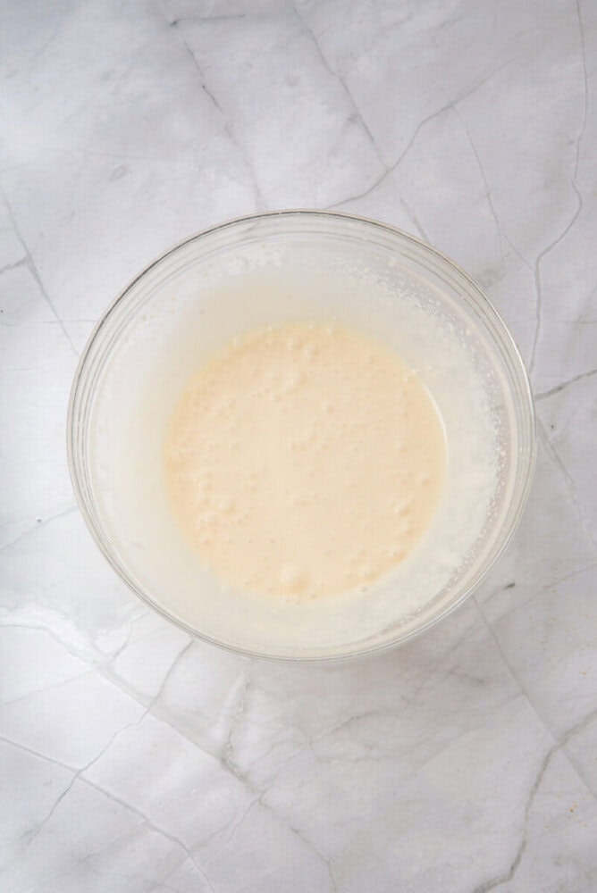 A glass mixing bowl containing a pale, slightly bubbly batter sits on a white marble surface.