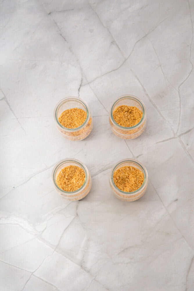 Four small glass jars filled with a tan, granular substance are arranged in a square on a light-colored marble surface.