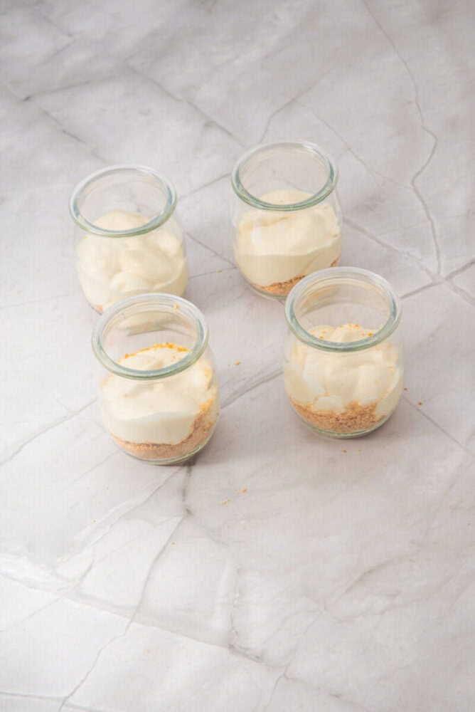 Four small glass jars with a layer of crushed graham crackers topped with creamy cheesecake filling, arranged on a light grey marble surface.