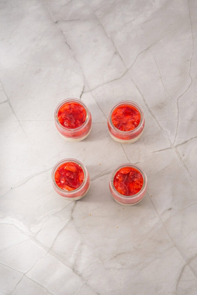 Four small glass jars filled with a red fruit dessert are arranged in a square pattern on a light marble surface.