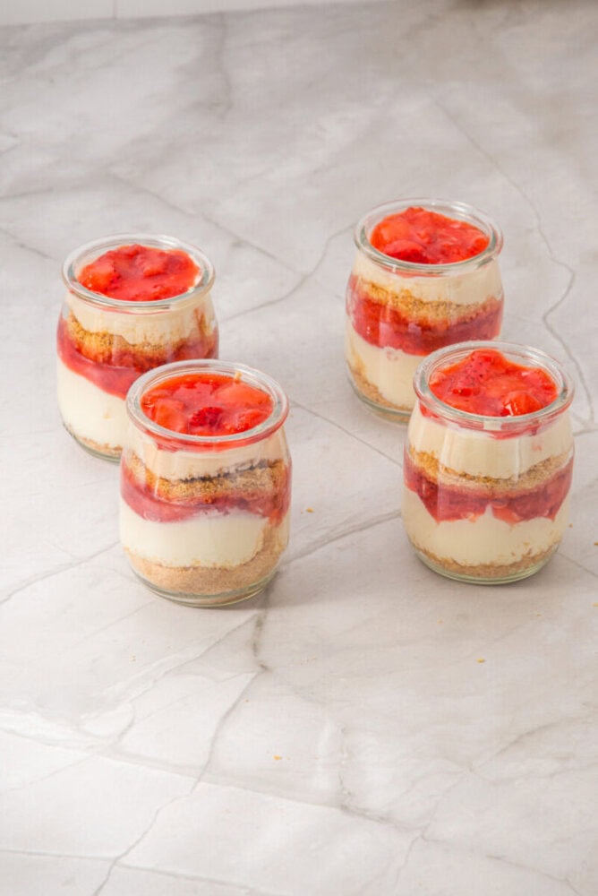 Four glass jars filled with layered cheesecake, strawberry sauce, and crumb crust are arranged on a light marble surface.