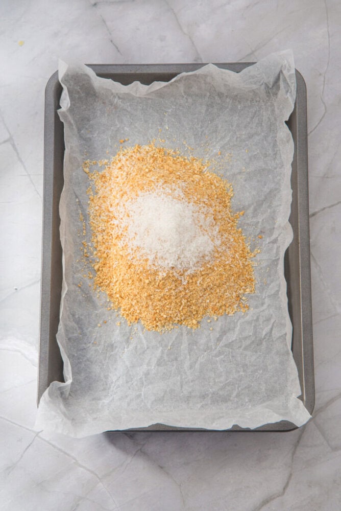 A baking tray lined with parchment paper holds a mound of breadcrumbs and grated Parmesan cheese.