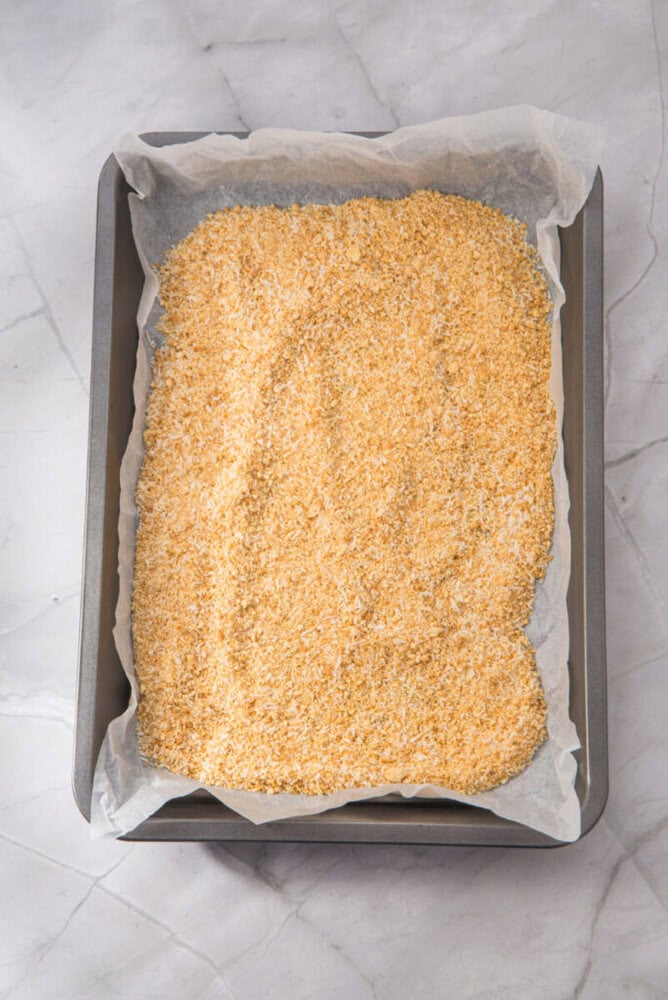 A rectangular baking tray lined with parchment paper and filled with an even layer of breadcrumbs on a marble surface.