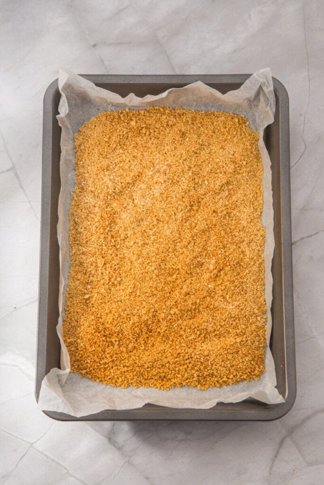 A baking tray lined with parchment paper holds an even layer of golden brown breadcrumbs on a marble surface.