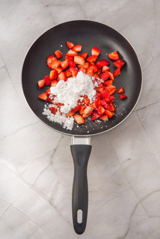 A nonstick frying pan with chopped strawberries and a pile of white sugar on a light marble countertop.