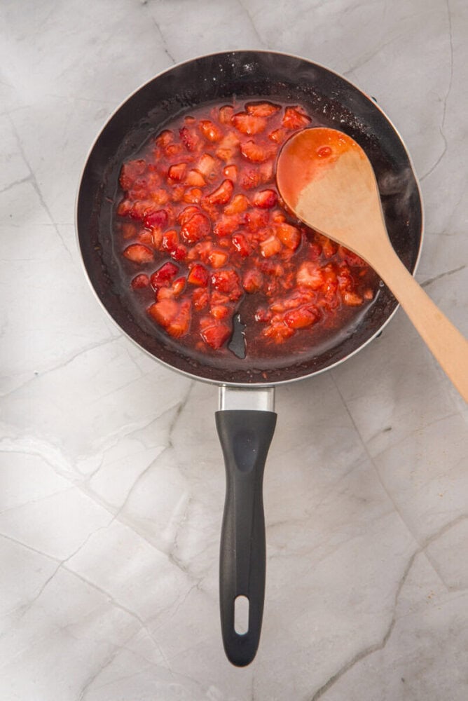 A frying pan with chopped strawberries cooking in syrup on a marble surface, stirred with a wooden spoon.