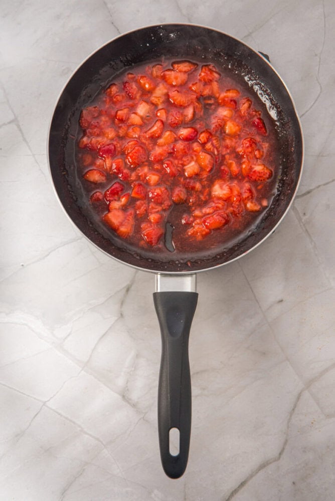 A black-handled frying pan with chopped strawberries cooking in syrup on a light-colored marble surface.