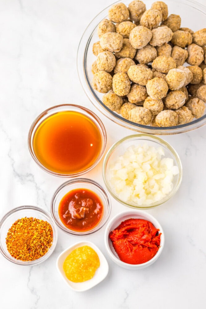 A bowl of meatballs beside bowls of hot sauce, diced onions, tomato paste, chili flakes, mustard, and chili sauce on a white surface.