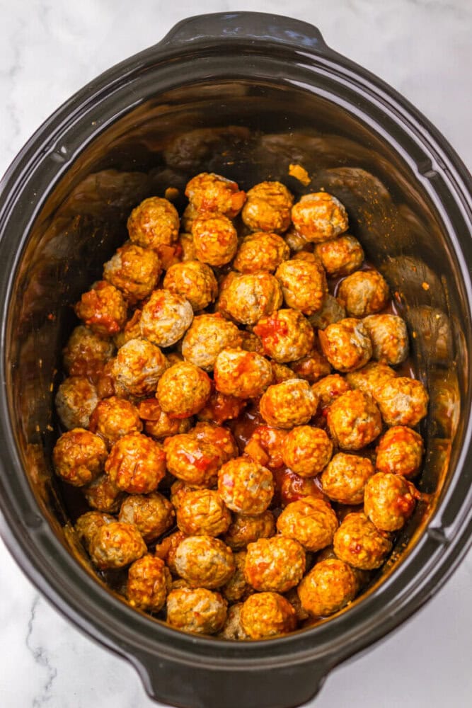 A slow cooker filled with meatballs coated in a red sauce, viewed from above on a marble surface.