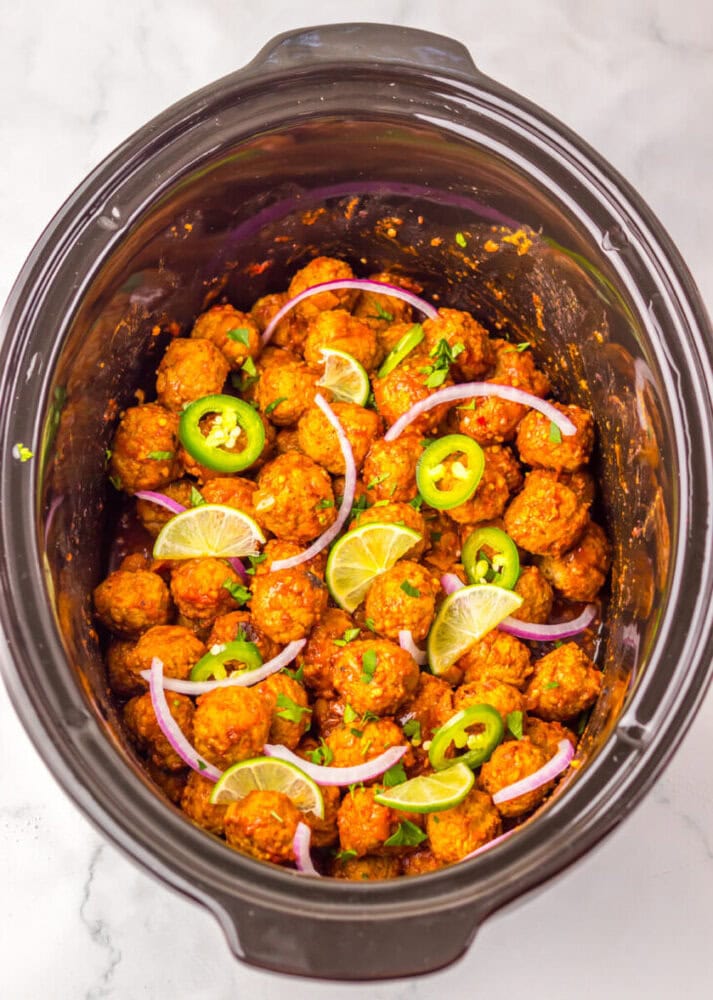 A slow cooker filled with seasoned meatballs, garnished with sliced lime, jalapeño, red onion, and chopped herbs.