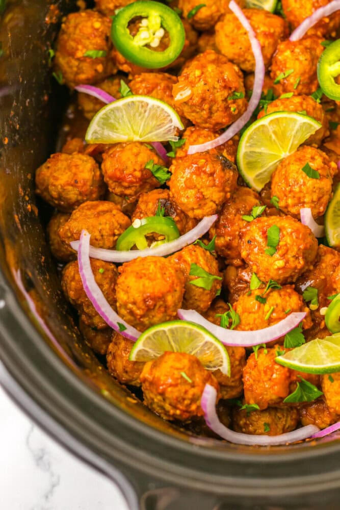 Slow cooker filled with seasoned meatballs garnished with lime slices, red onion, jalapeño slices, and chopped herbs.