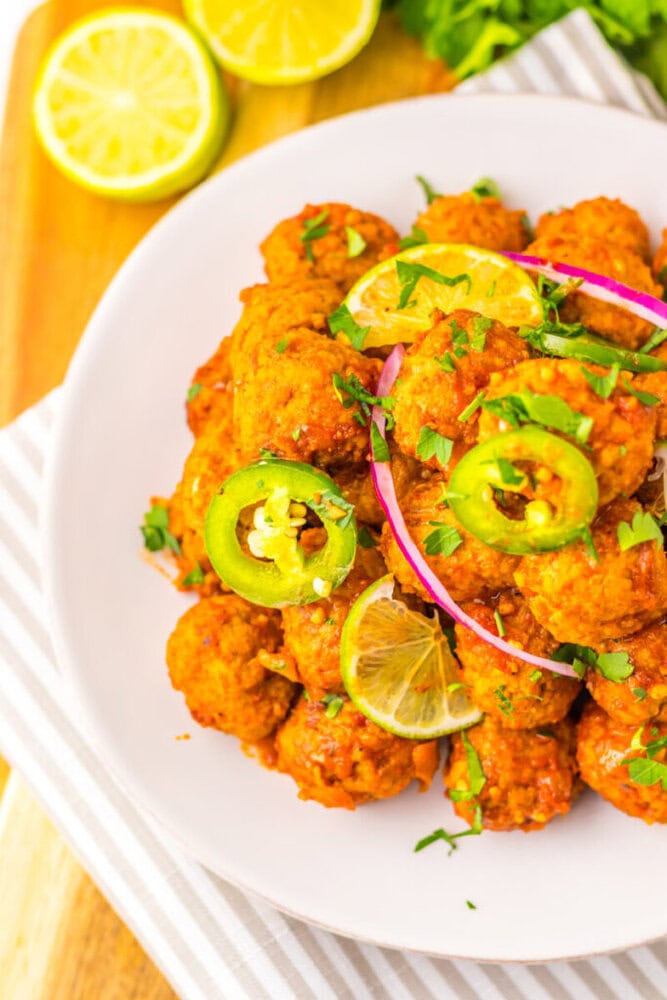 A white plate of spicy fried meatballs garnished with sliced jalapeños, red onion, chopped herbs, and lemon slices, placed on a striped napkin.