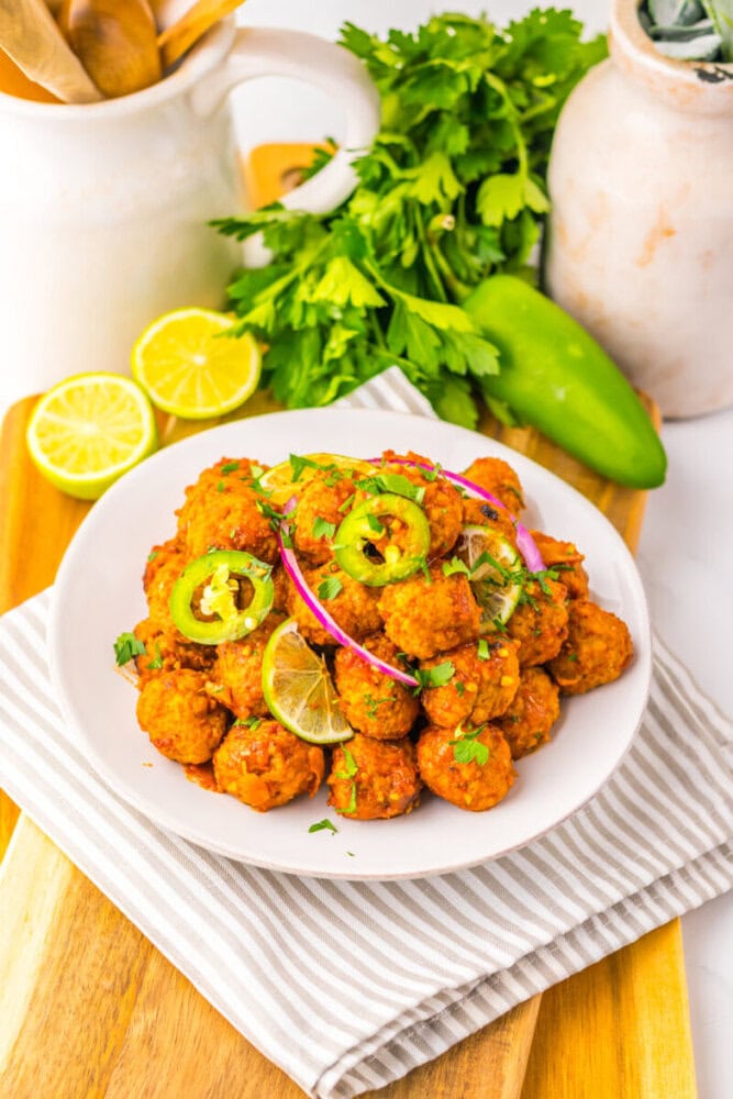 A white plate of fried vegetable balls garnished with sliced jalapeños, red onion, lime, and cilantro, set on a striped napkin with herbs and lemon in the background.
