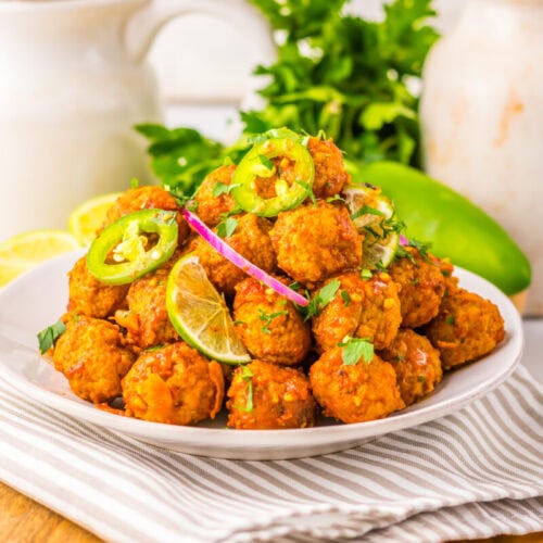 A plate of fried vegetarian meatballs garnished with lime slices, jalapeño, and herbs, placed on a striped cloth with ingredients in the background.