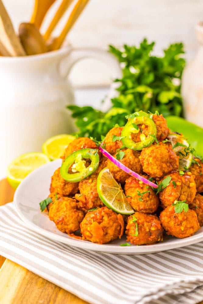 A white plate of spicy vegetarian meatballs garnished with lime wedges, jalapeño slices, red onion, and herbs sits on a striped napkin with fresh parsley and lemons in the background.