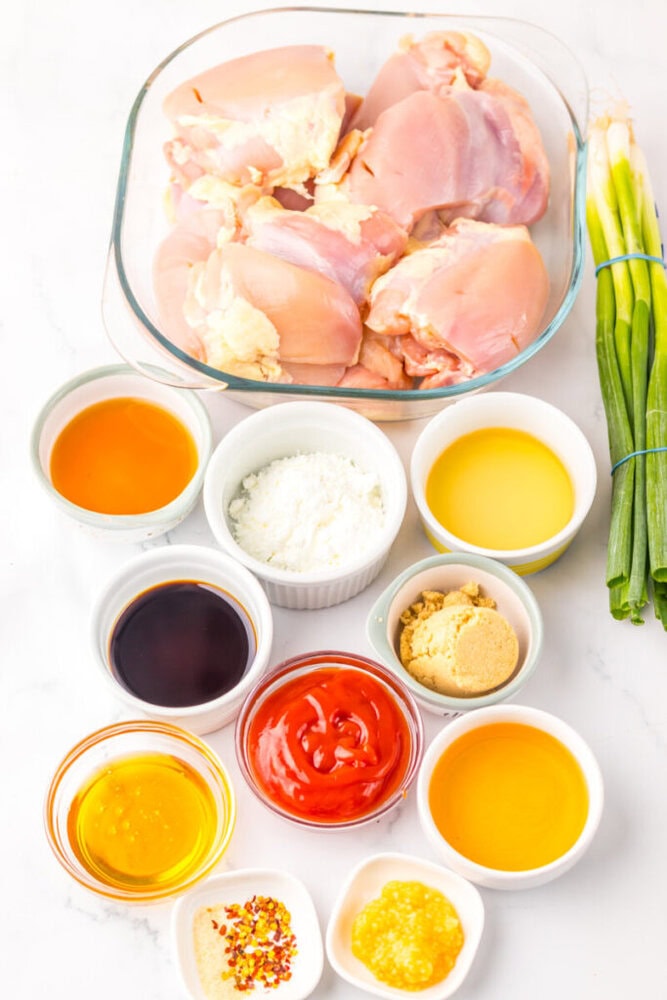 Raw chicken thighs in a glass dish surrounded by small bowls containing various sauces, seasonings, cornstarch, brown sugar, and green onions on a white surface.
