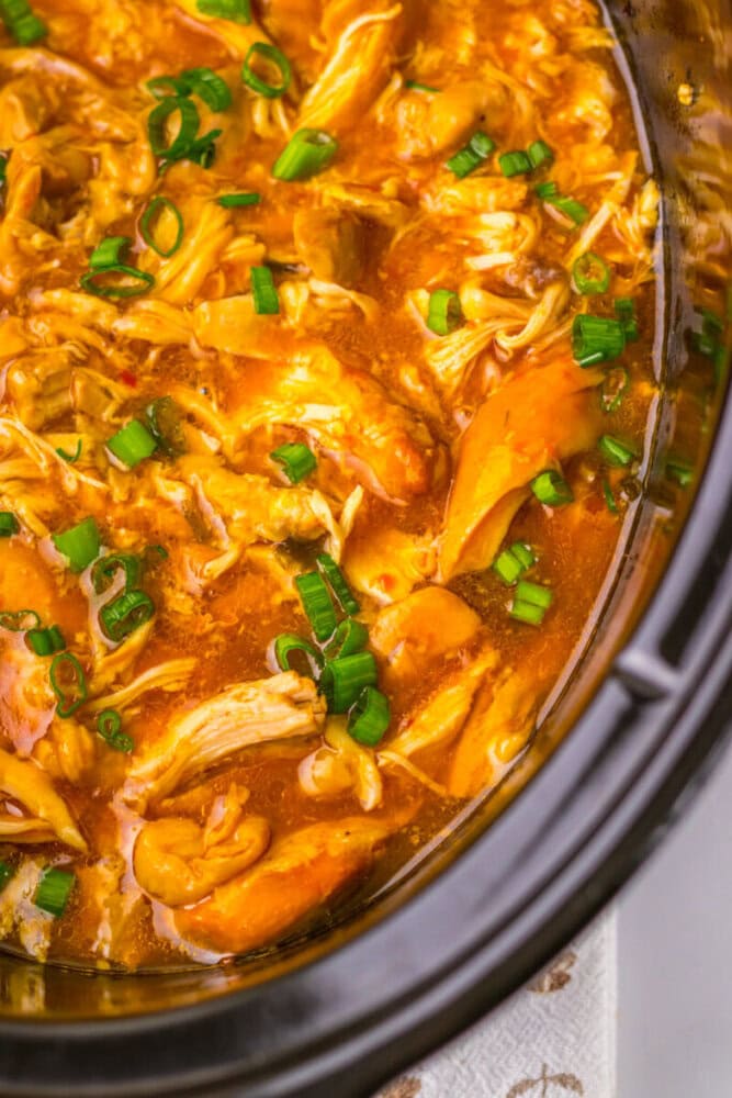 Shredded chicken in a thick orange sauce garnished with chopped green onions, served in a slow cooker.