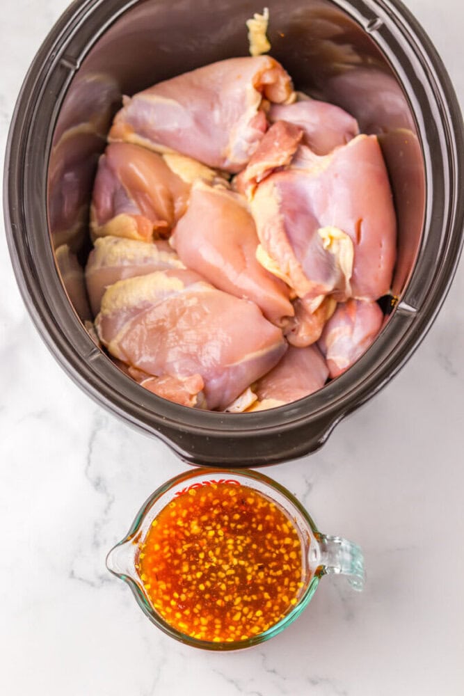 Raw chicken thighs arranged in a slow cooker, with a glass measuring cup of orange-colored sauce containing visible chili flakes beside it on a white countertop.
