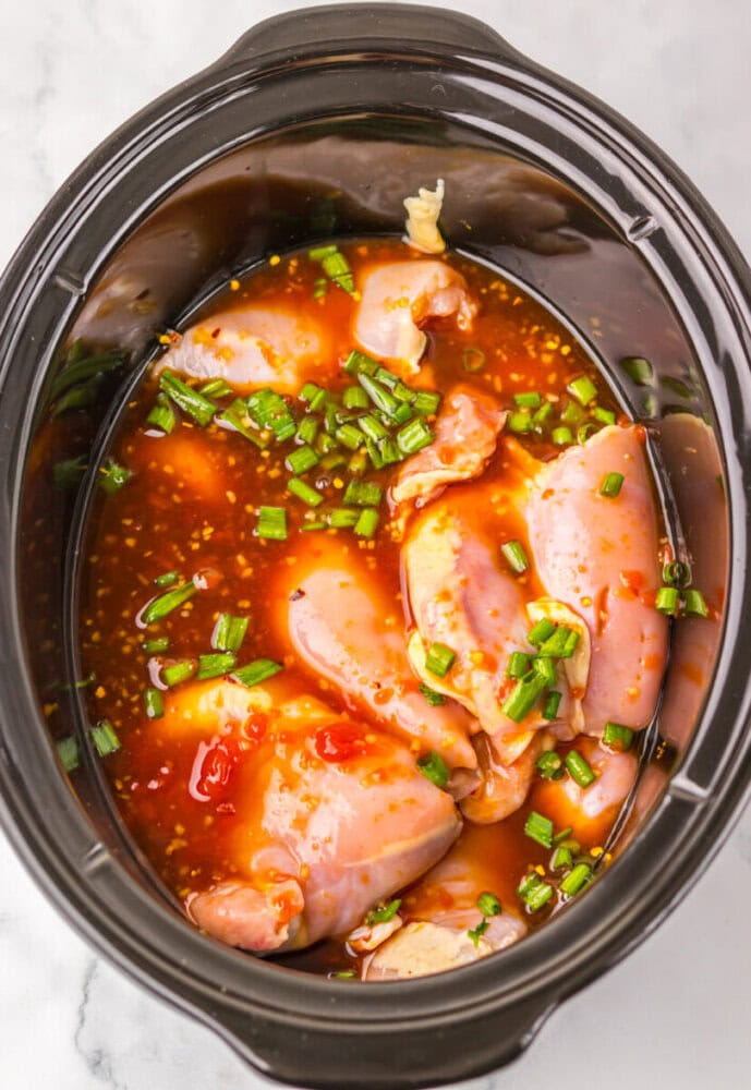 Raw chicken thighs in a slow cooker with a red sauce and chopped green onions on top, ready to be cooked.