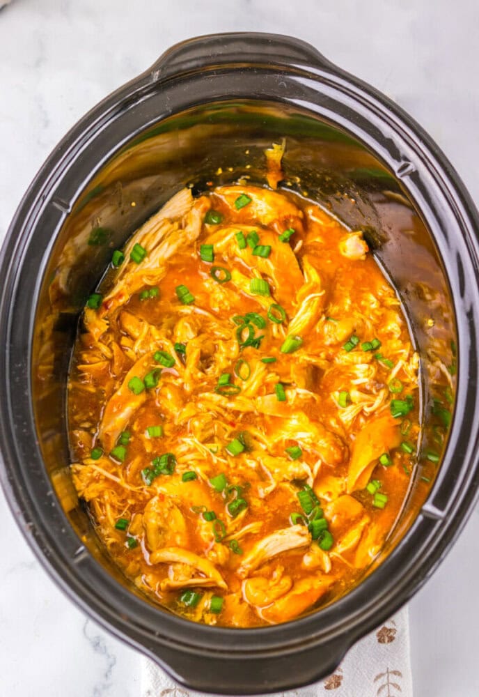 Shredded chicken in a slow cooker with a thick orange sauce, topped with chopped green onions.