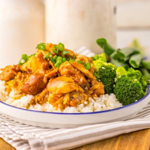 A plate of white rice topped with a saucy meat and vegetable stew, garnished with chopped green onions, served with broccoli and leafy greens on the side.